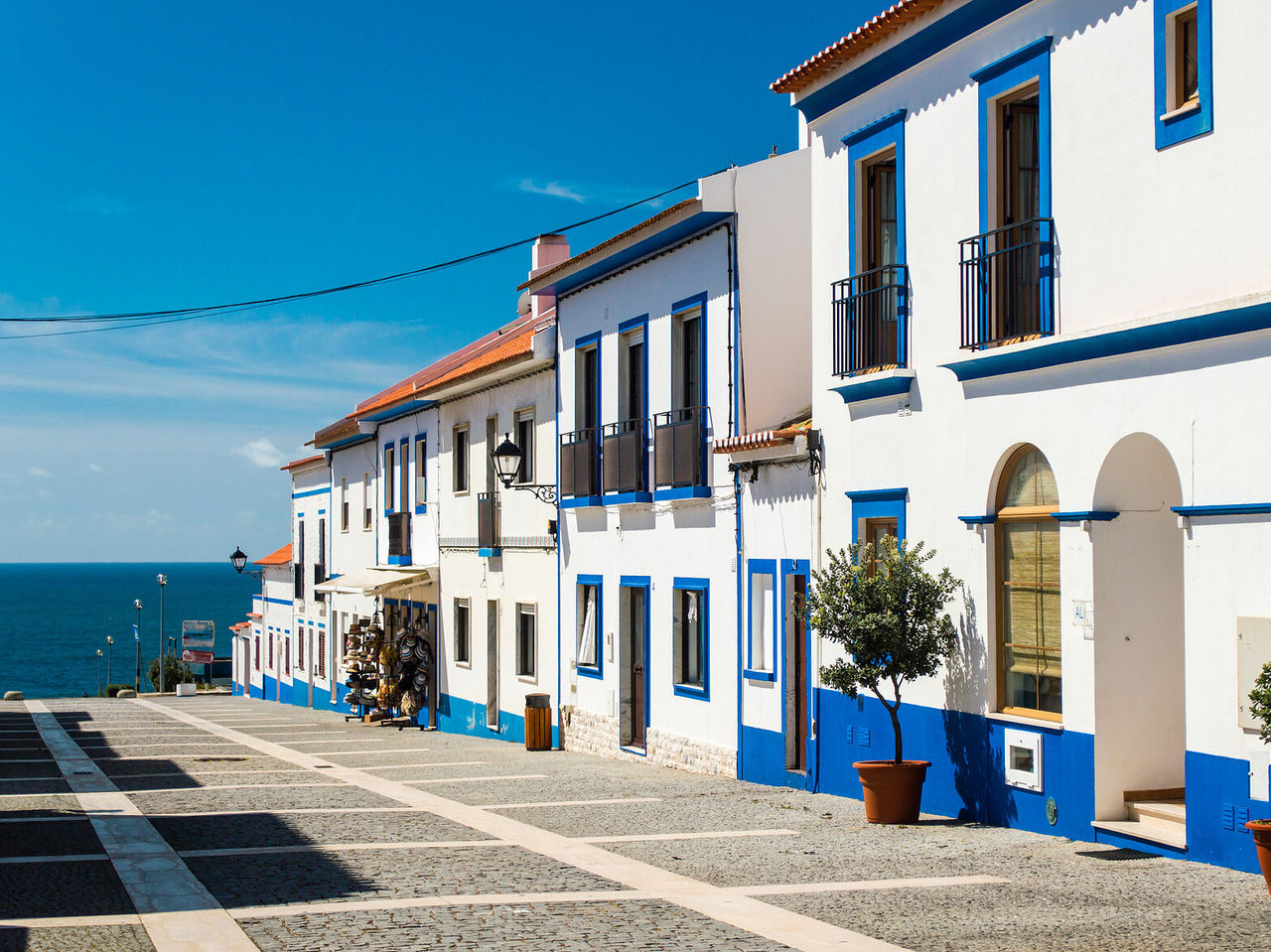 Calle con casas típicas de la región del Alentejo, blancas y azules con macetas en la puerta, y una vista increíble al océano