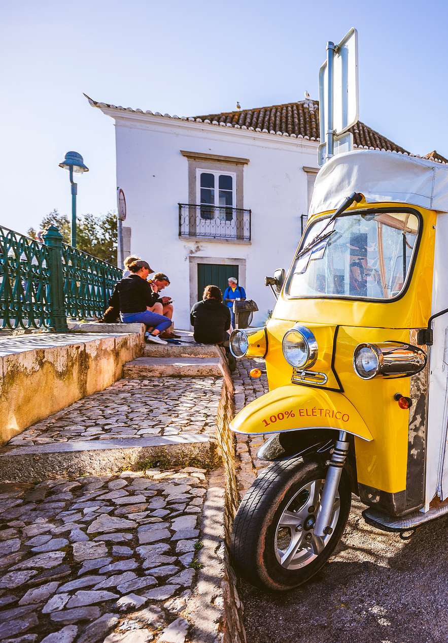 Tours en TukTuk en Algarve, con un TukTuk amarillo en el centro de la ciudad y gente socializando al fondo