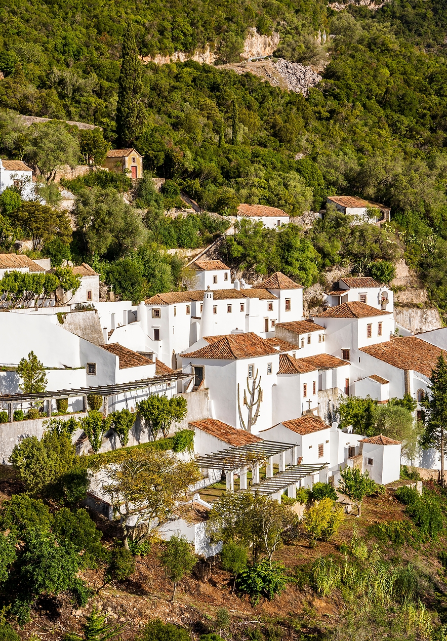 Convento da Serra da Arrábida, com vários edifícios brancos, rodeado de paisagem montanhosa e vegetação densa