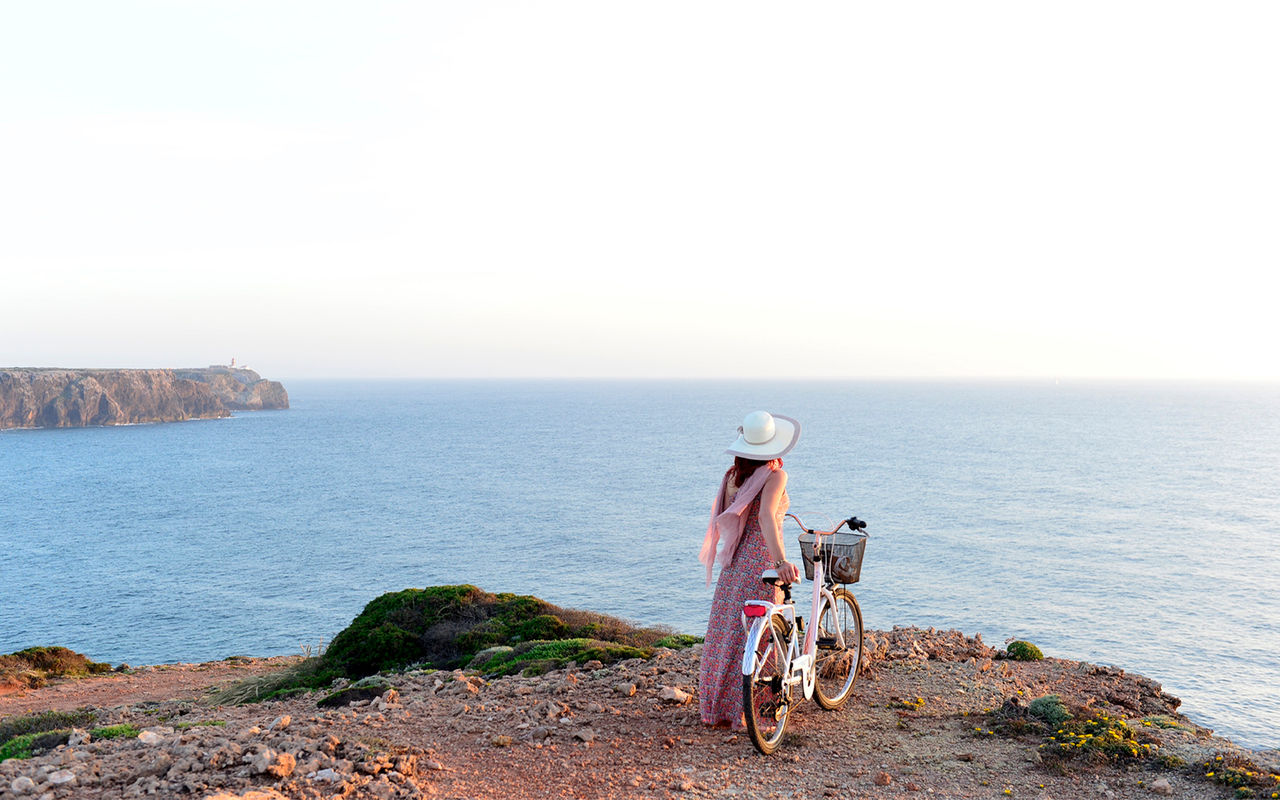 Pessoa com bicicleta observa o mar sobre falésias rochosas ao entardecer sob céu claro e horizonte aberto
