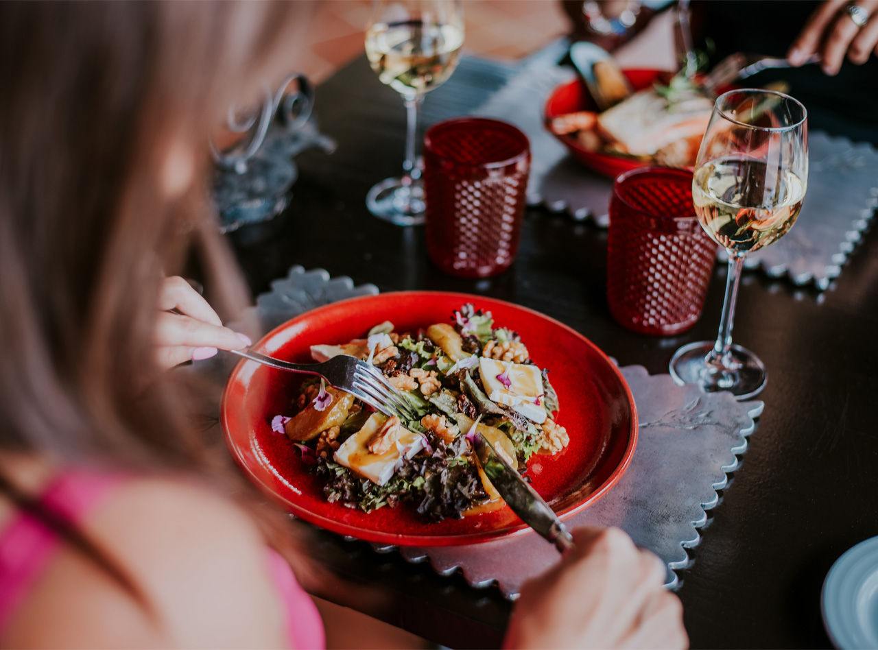 Pessoa desfrutando salada servida em prato vermelho sobre mesa escura com taças de vinho e velas decorativas