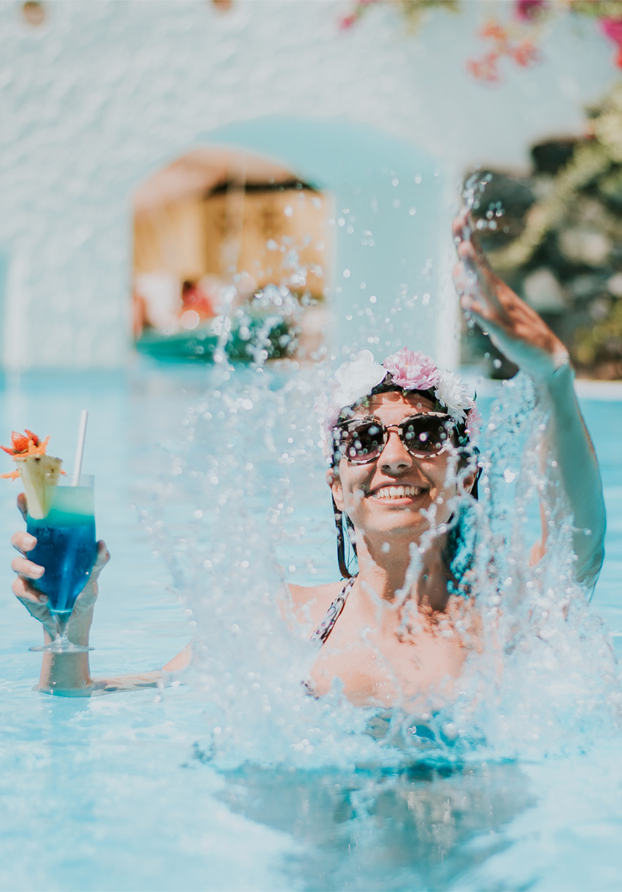 Frau mit Sonnenbrille und Cocktail spritzt lachend im türkisblauen Pool vor weißen Bögen und Pflanzen