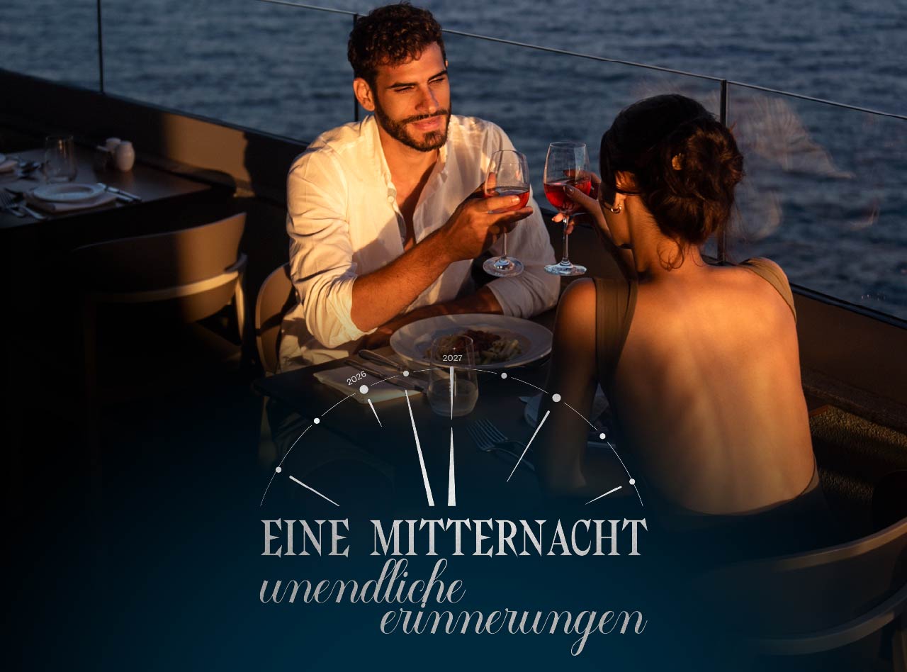 Paar beim Abendessen mit Rotweingläsern auf einer Terrasse am Meer im warmen Licht der Abendsonne am Fenster