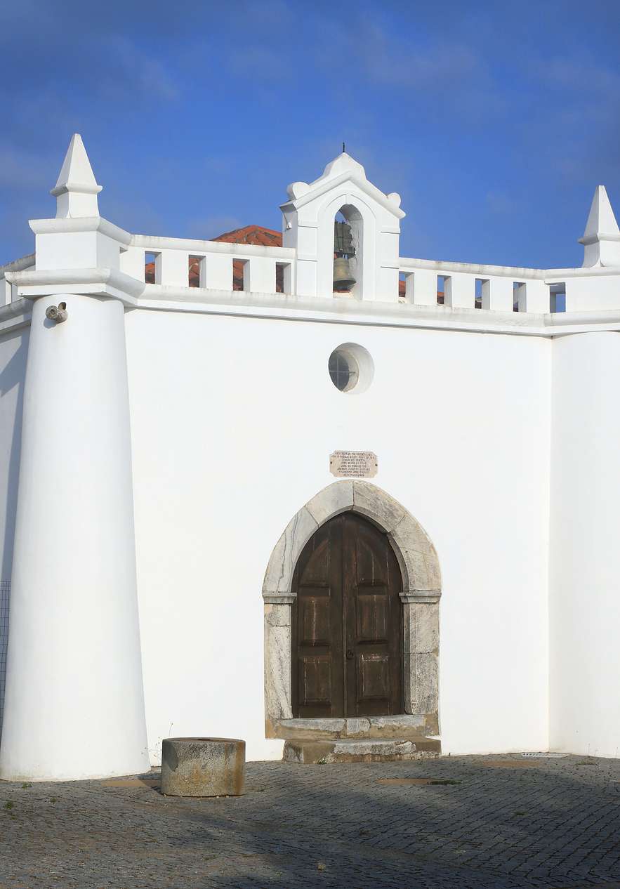 White convent facade with arched wooden doorway, bell tower and cobbled square beneath a bright blue sky