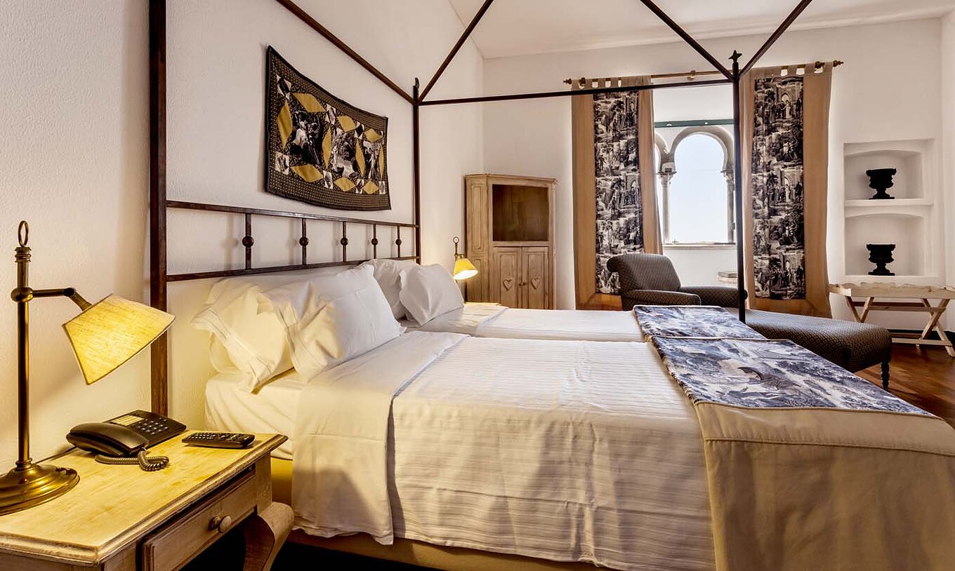 The Standard Suite of the Pousada Castelo Alvito has two beds, two bedside tables, an armchair, and a window