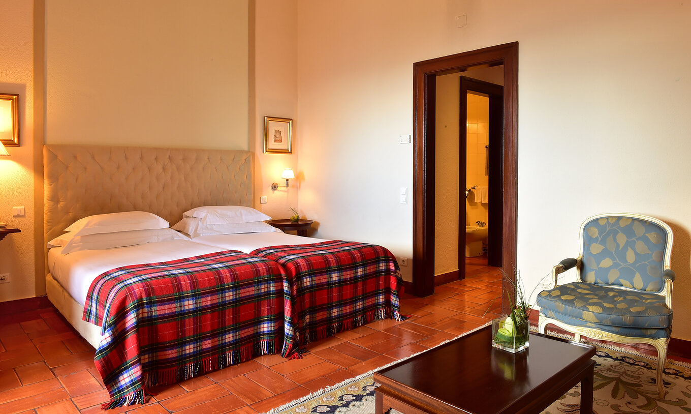 The Superior Family Room at Pousada Castelo Palmela has two beds with a red checkered blanket, bedside table, and armchair