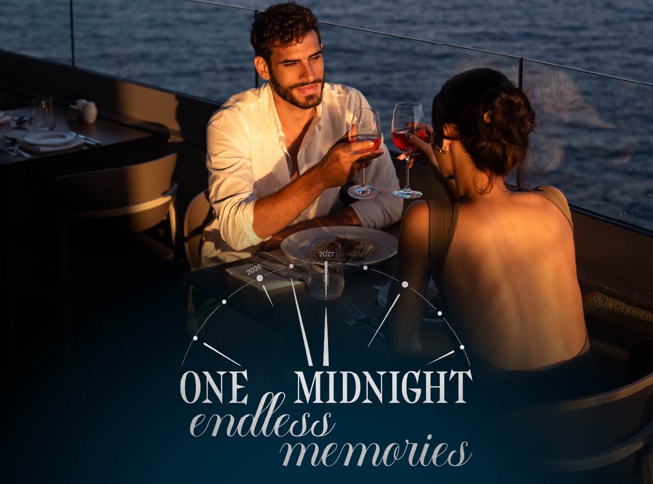 Couple raising wine glasses over dinner beside the sea at sunset with One Midnight endless memories lettering