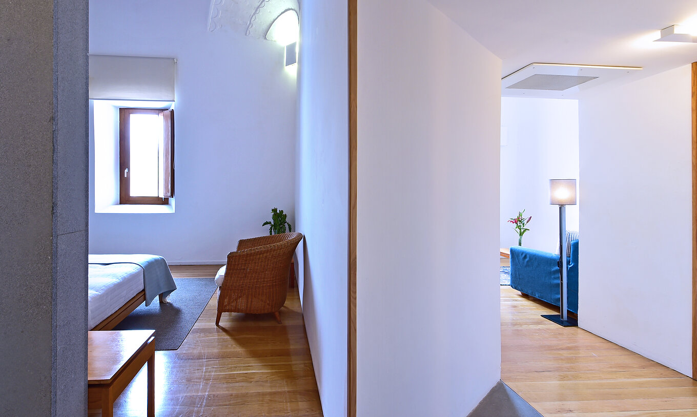 The Standard Suite of The Pousada Convento Arraiolos has a room with a bed and an armchair and a hall with a sofa
