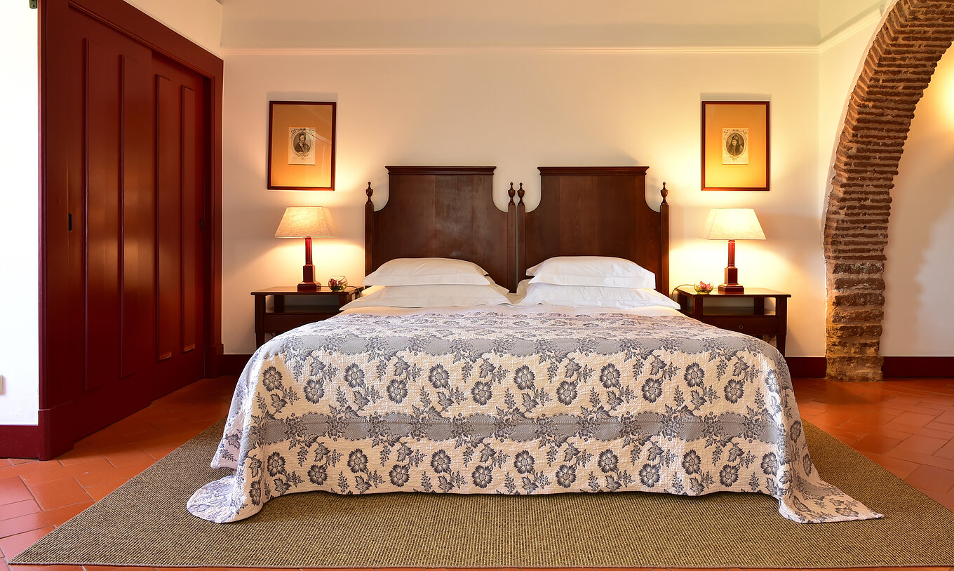 The Junior Suite of The Pousada Convento Beja has a double bed with a blue bedspread, a wardrobe, and two bedside tables