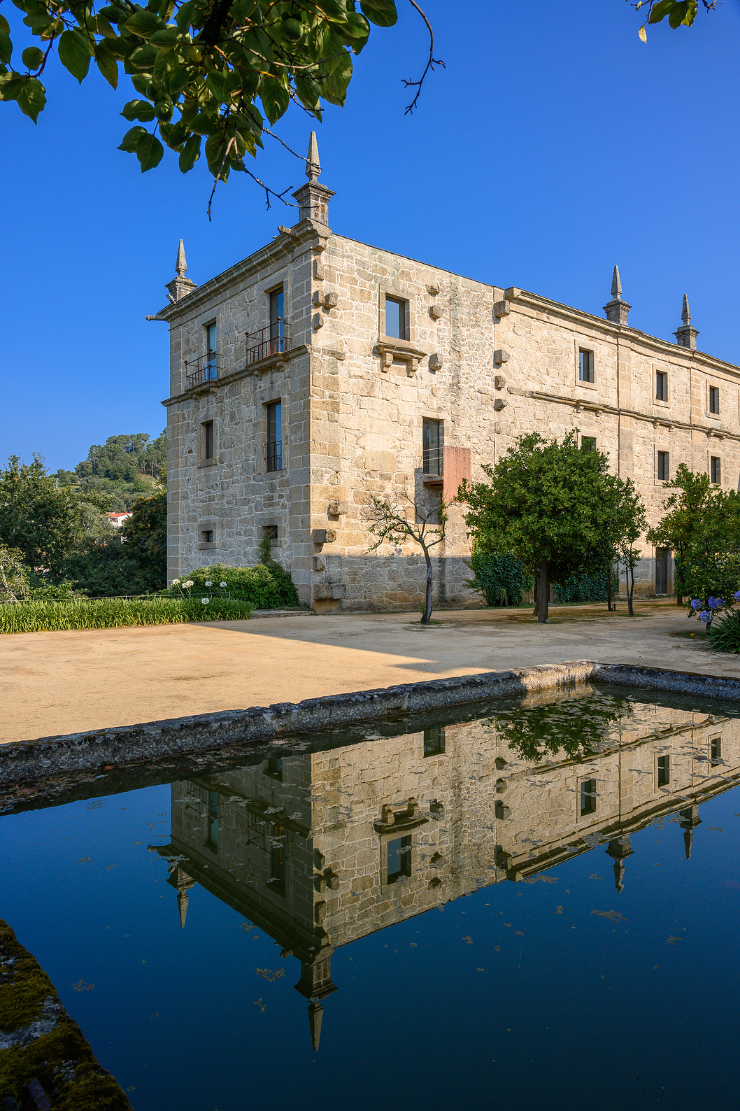 Side facade of Pousada Mosteiro Amares, with a stone wall, a pond in front, and trees