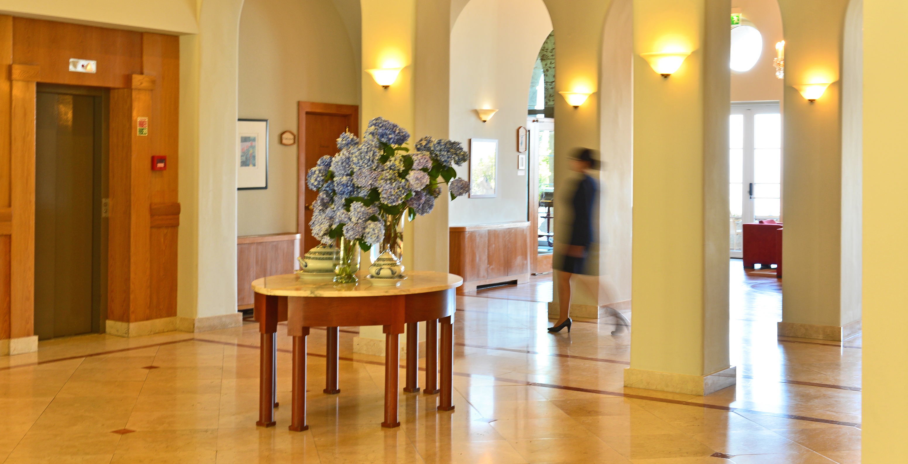Entrance hall with a table in the center with flower vases and a lady passing in the background