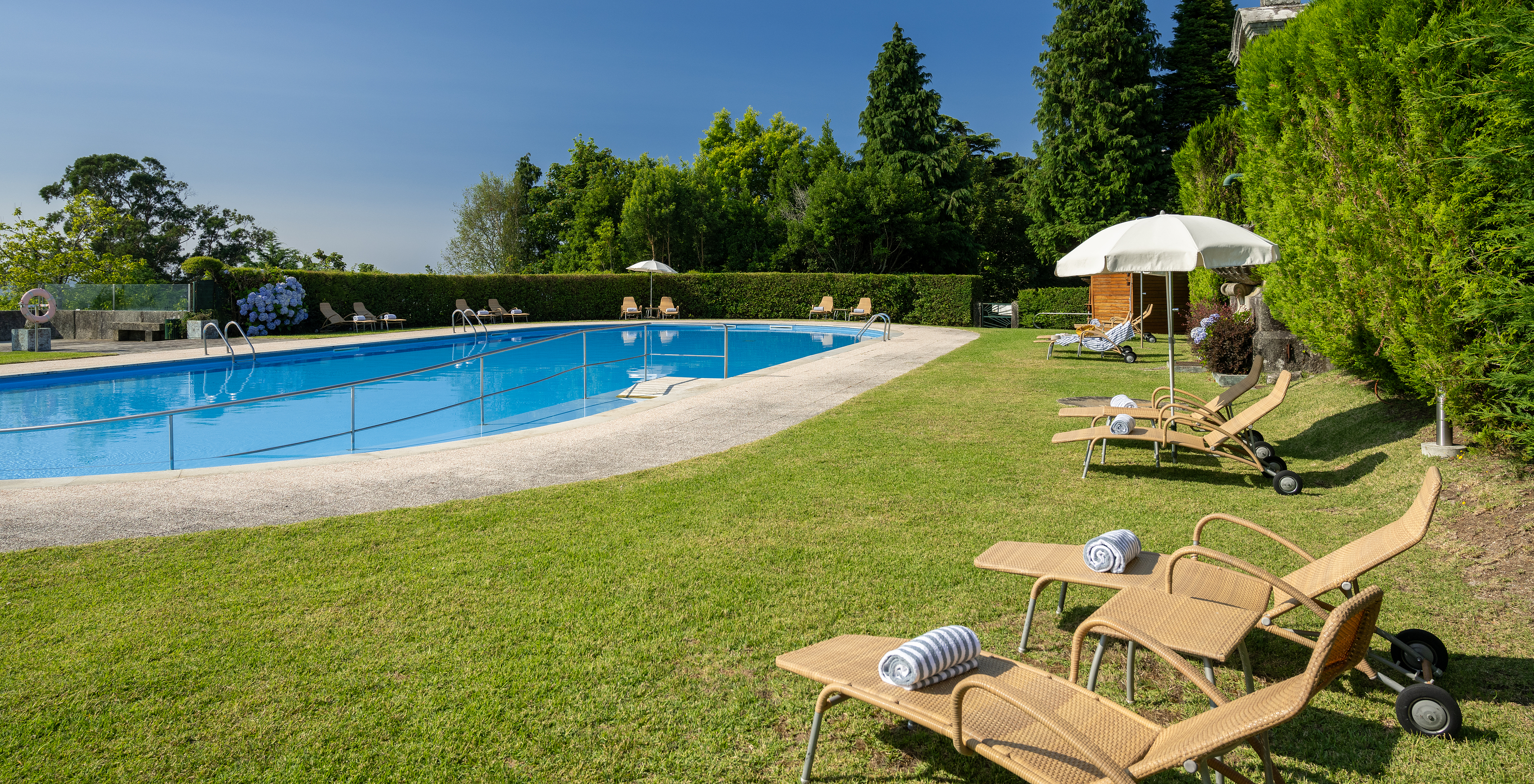 Outdoor pool with sun loungers and umbrellas, with green gardens and several trees in the Pousada Viana do Castelo