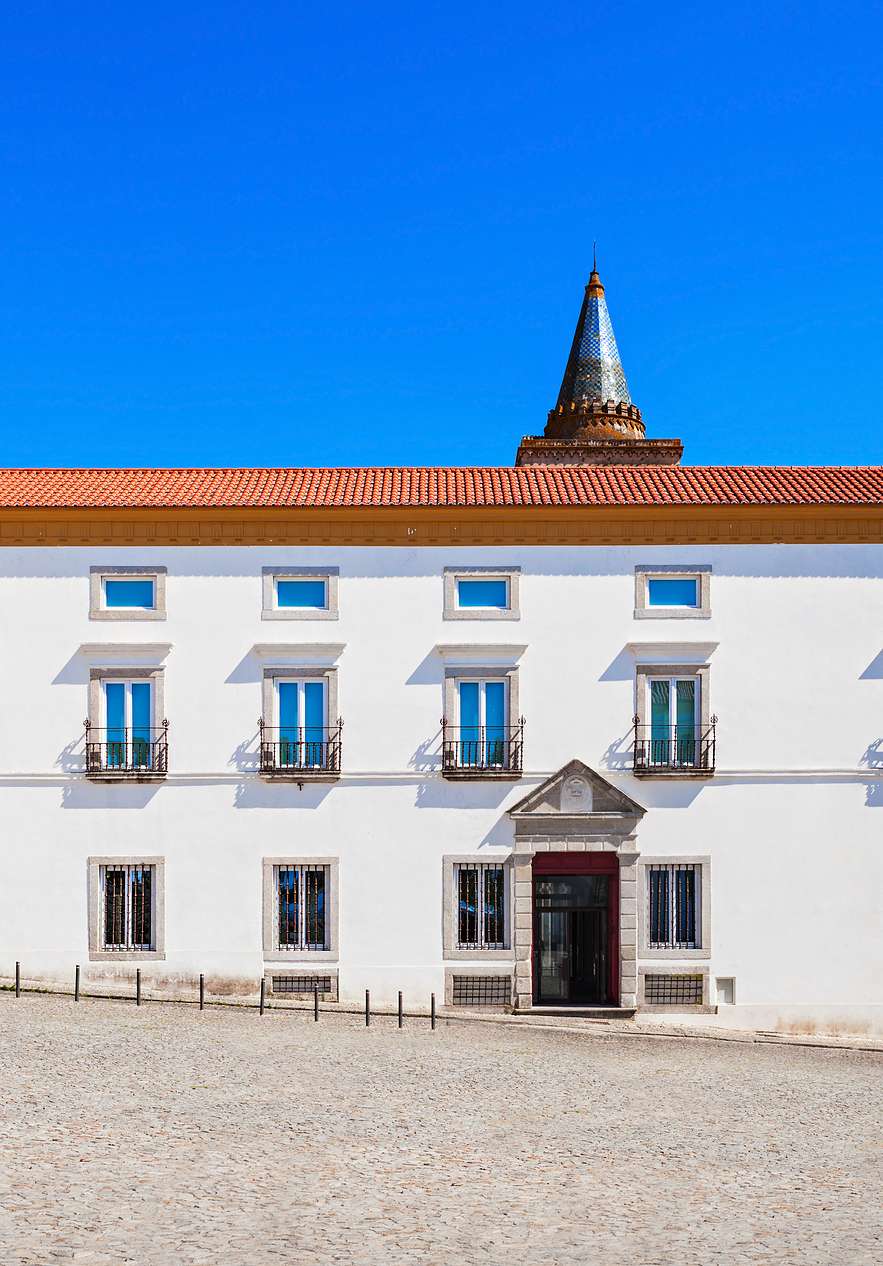 Fachada blanca con ventanas simétricas, tejado de tejas y torre histórica bajo un cielo azul intenso