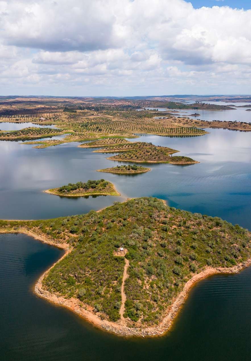 Vista aérea de un embalse con pequeñas islas cubiertas de vegetación y colinas bajo nubes dispersas