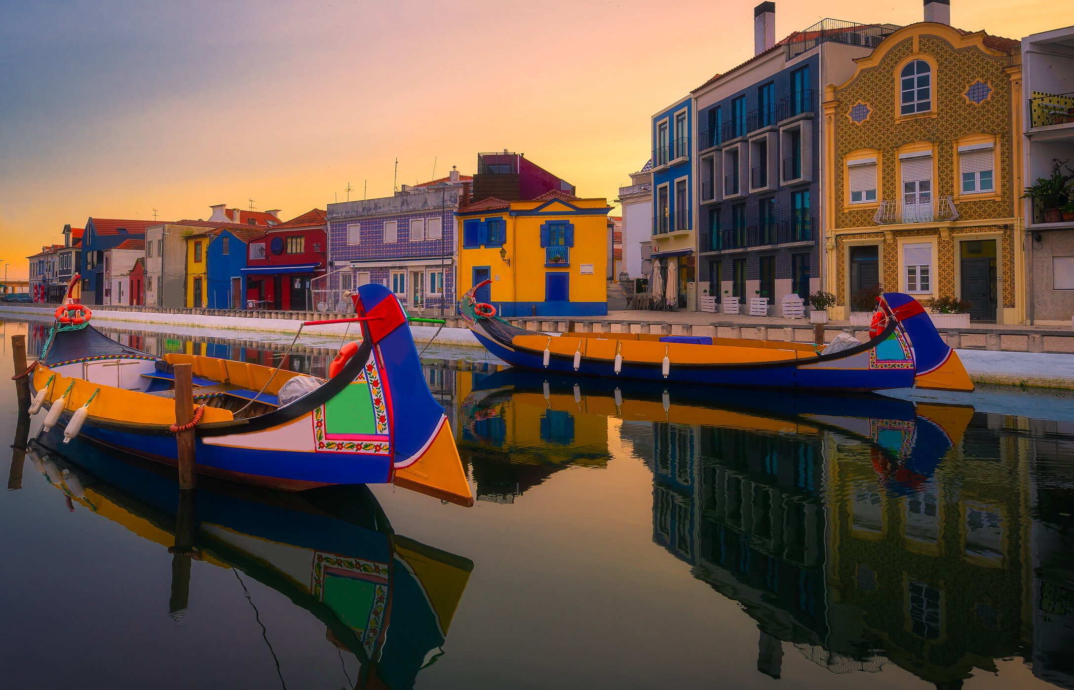 Vista de dos moliceiros anclados, típicos de la región de Aveiro en Portugal, con edificios tradicionales al fondo.