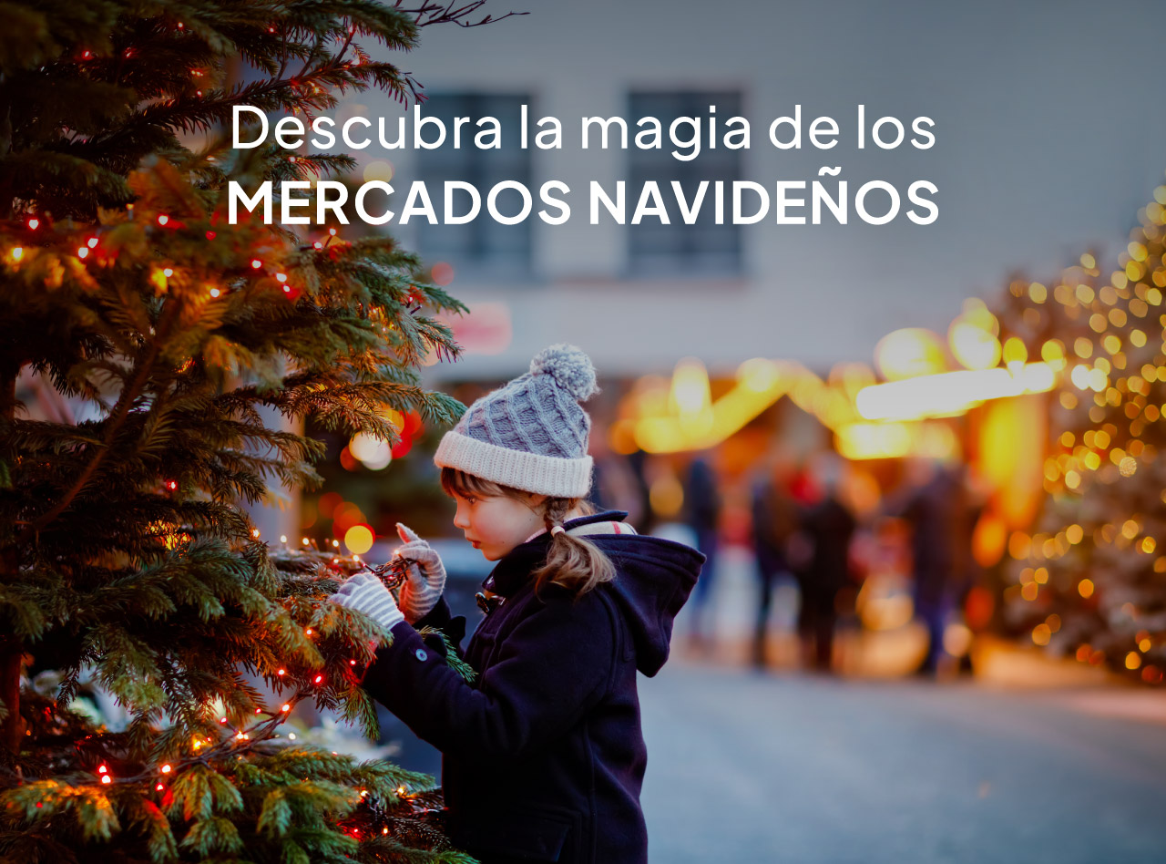 Niña con gorro de invierno decorando un árbol iluminado en un mercado navideño con luces desenfocadas