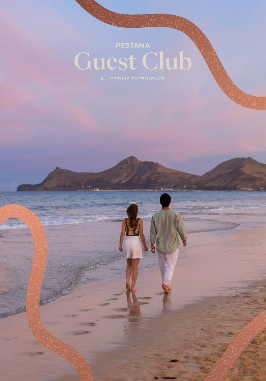 Pareja caminando por la orilla al atardecer con montañas frente al mar y cielo rosado sobre la playa