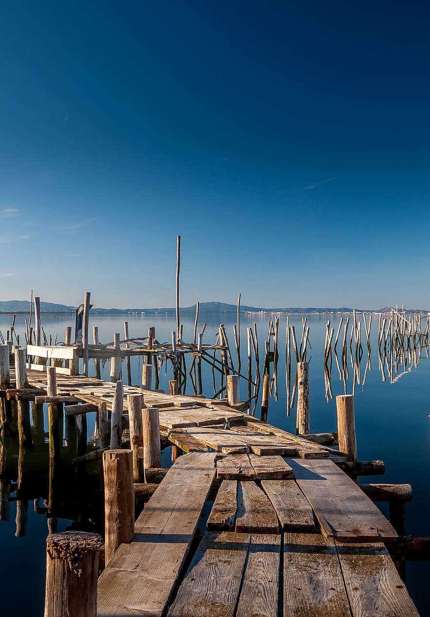 Ponton palafittique en bois de Carrasqueira avançant sur une eau calme sous un vaste ciel bleu et des pilotis reflétés