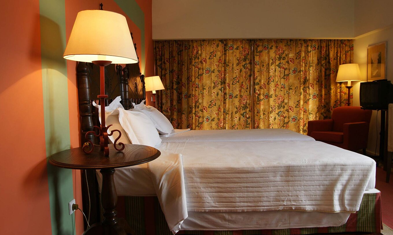 La Junior Suite de la Pousada Valença a deux lits séparés, une table de chevet, un fauteuil et un rideau à fleurs