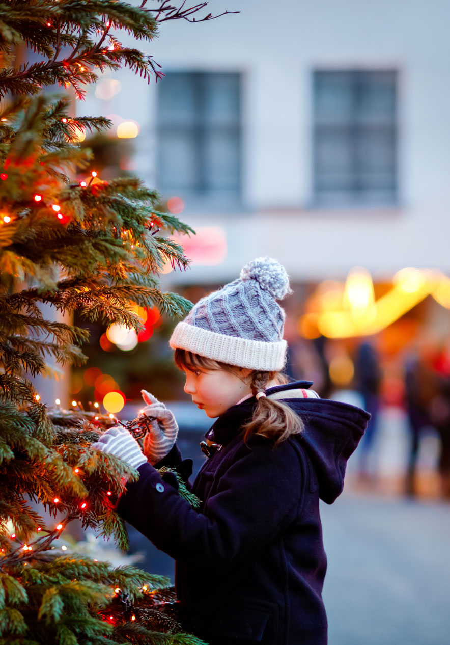 Enfant en bonnet gris décorant un sapin illuminé devant un marché de Noël aux lumières floues au crépuscule