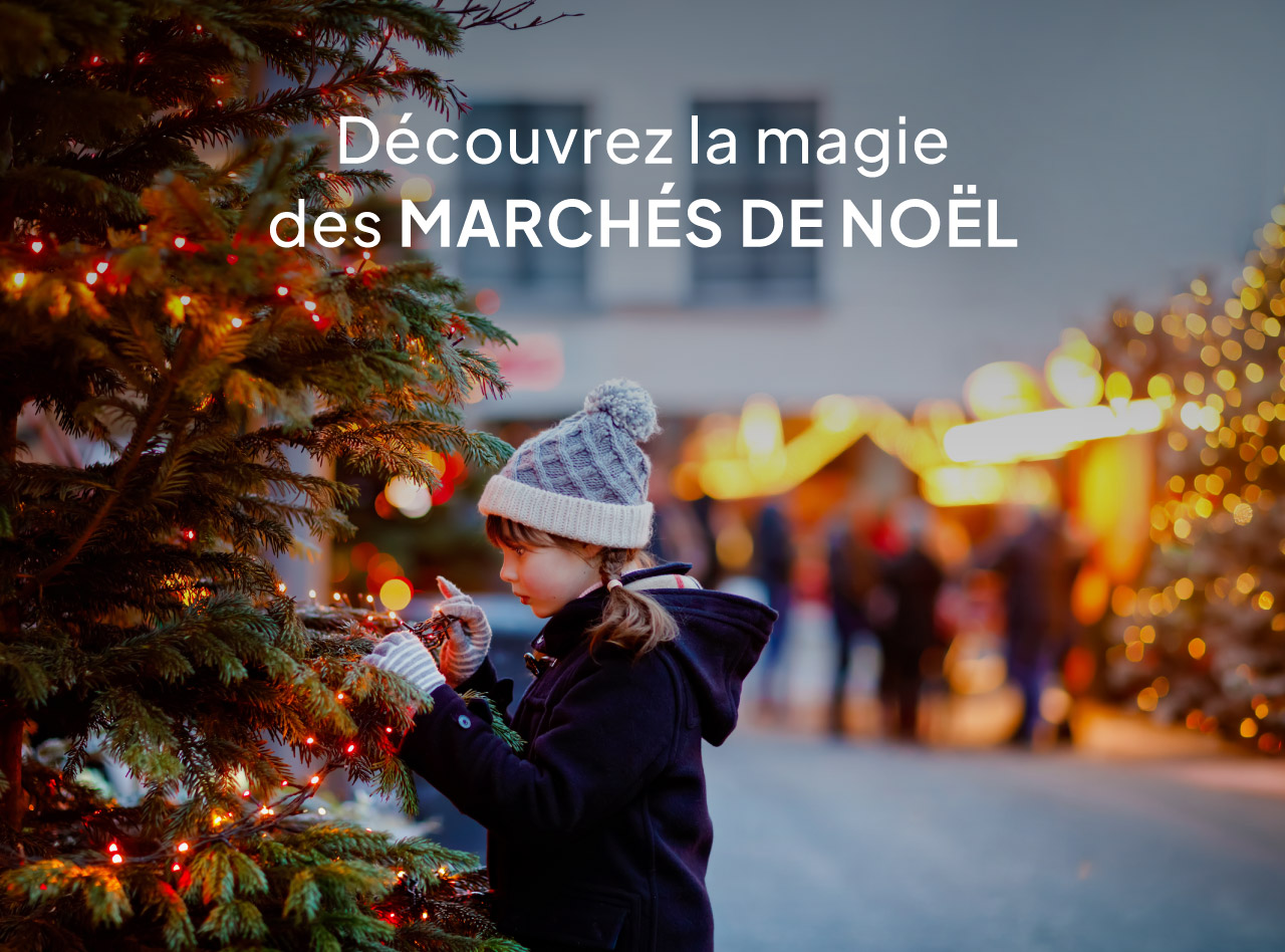 Enfant en bonnet gris décorant un sapin illuminé devant un marché de Noël aux lumières chaleureuses