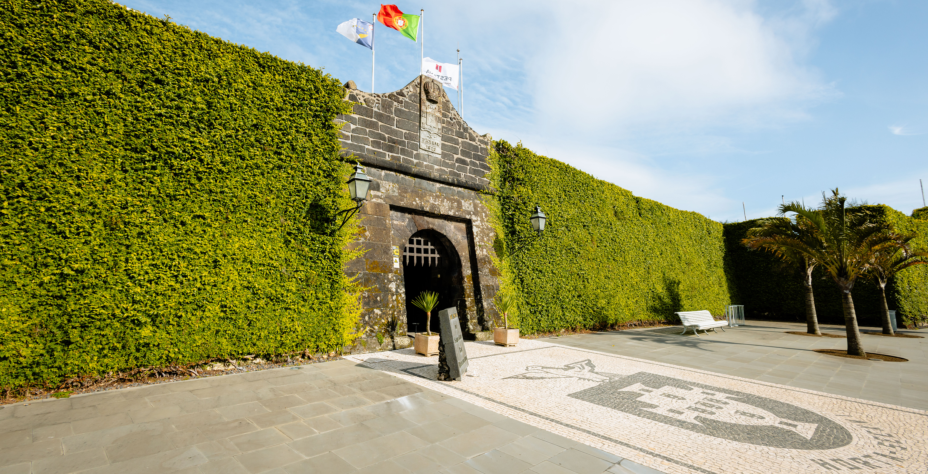 Fachada da entrada medieval da Pousada Forte Horta, com longas muralhas revestidas com densa vegetação e bandeiras