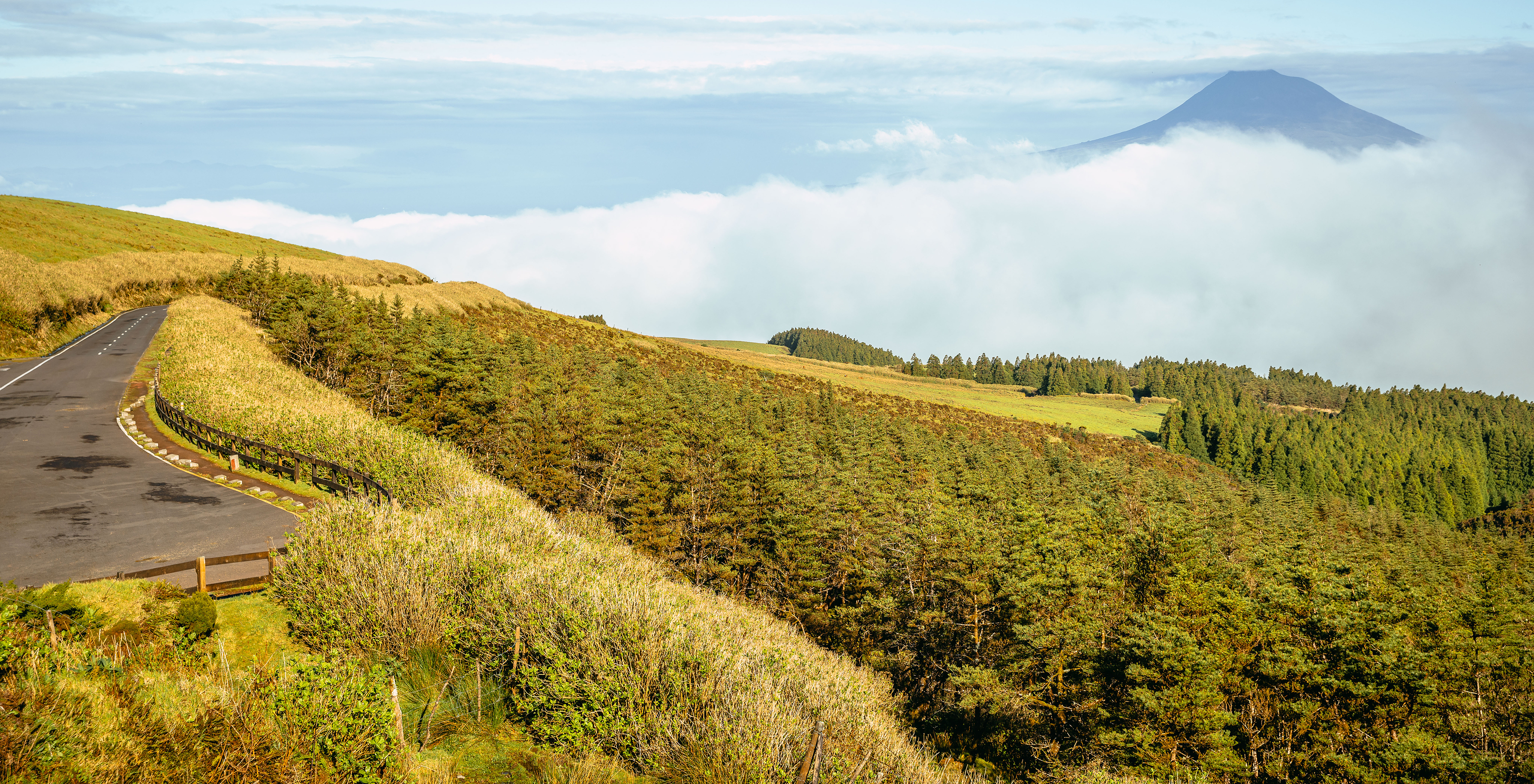 Vista panorâmica de uma estrada rural na Ilha do Faial, com um vulcão no horizonte, parcialmente encoberto por nuvens