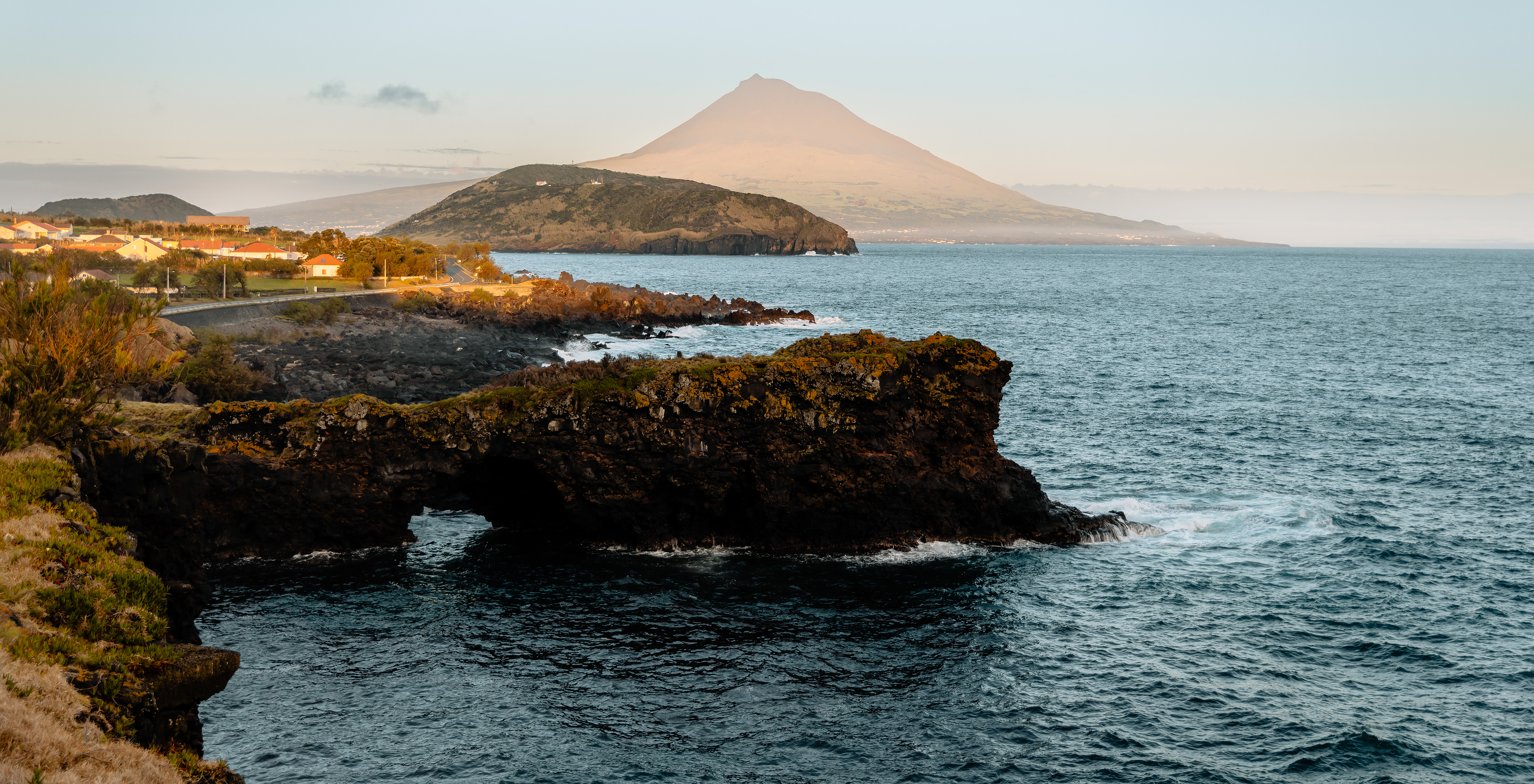Paisagem costeira exuberante nos Açores, com um vulcão no horizonte, uma pequena vila à beira-mar e rochas vulcânicas