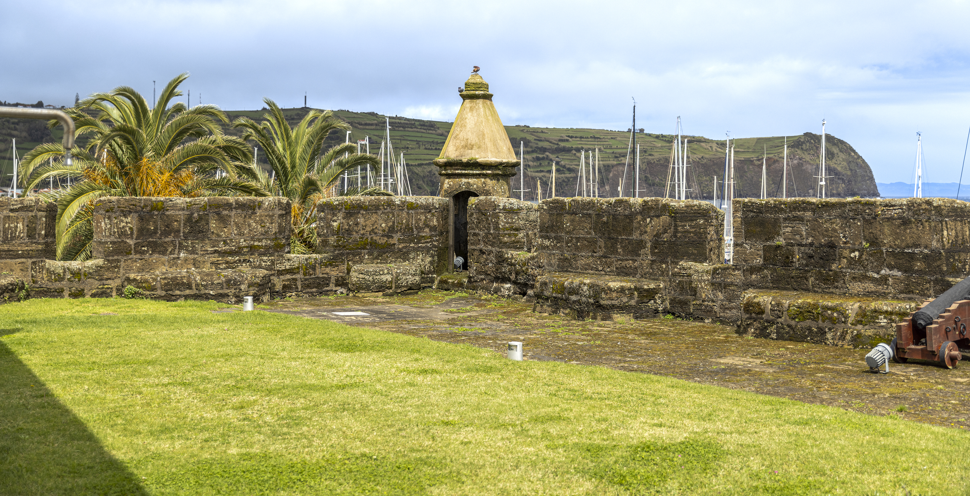 Vista de um forte histórico com muralhas de pedra, um canhão antigo, palmeiras e um mirante com vista para o mar