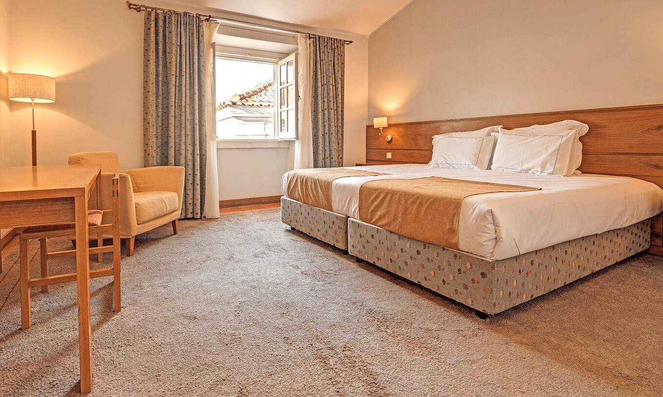 O Quarto Superior Família da Pousada Ourém tem duas camas juntas com uma janela ao lado aberta com vista para outras casas