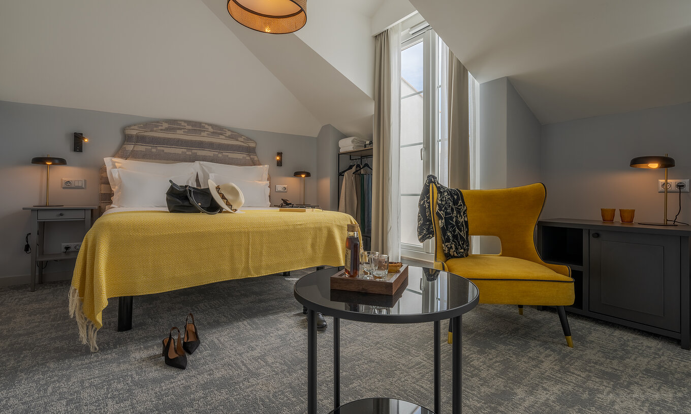 A Junior Suite da Pousada Alfama tem uma cama de casal com uma manta amarela, uma pequena mesa e um cadeirão amarelo