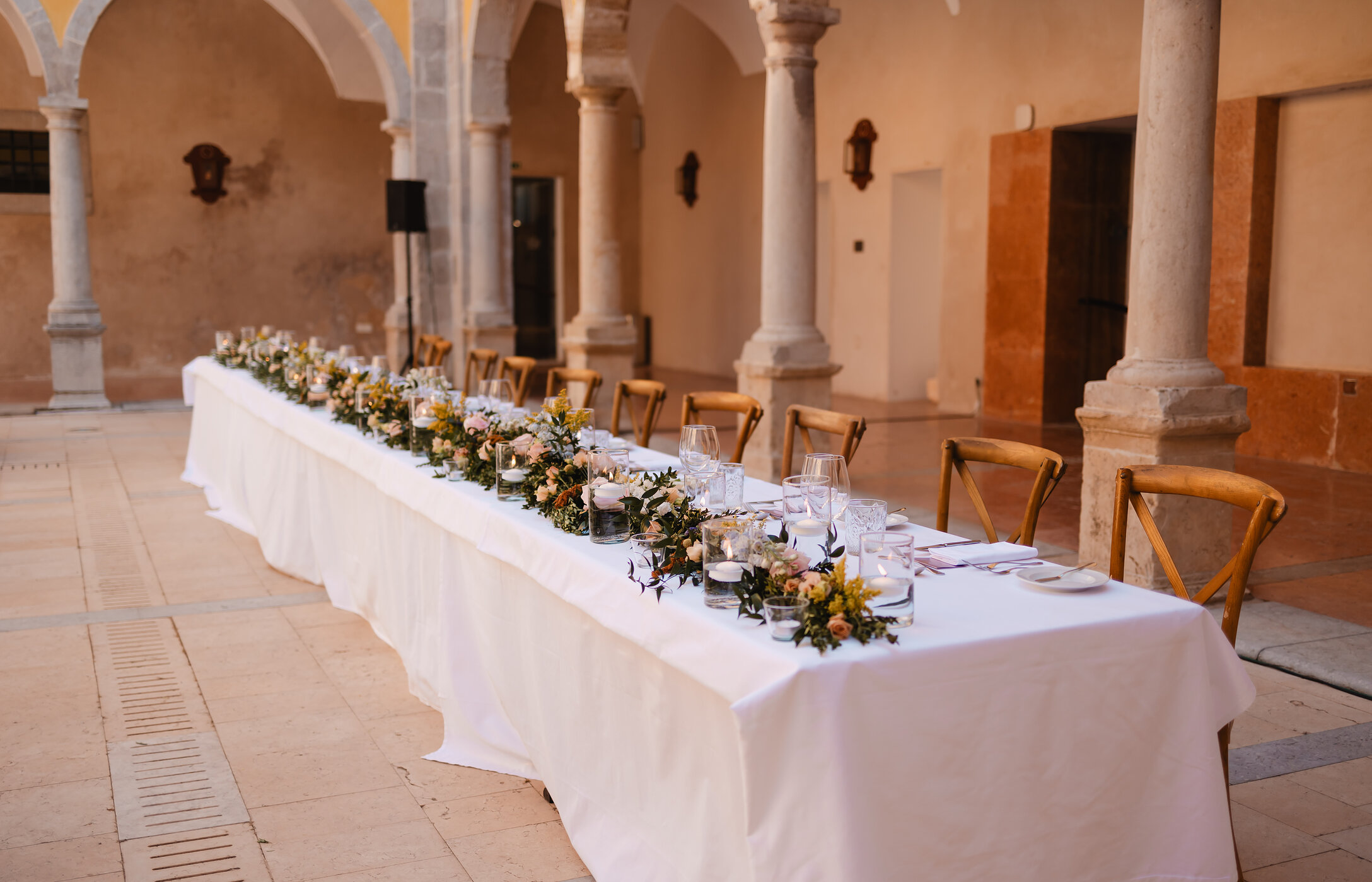 Mesa de noivos em pátio histórico com arcos e colunas, com decoração elegante e arranjos florais e cadeiras de madeira