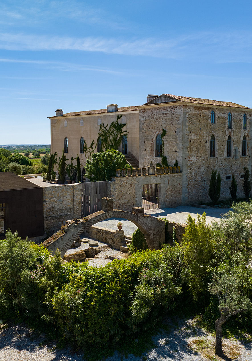 Vista exterior da Pousada Convento Arraiolos com fachada de pedra coberta por heras, rodeada por vegetação e campos sob céu azul