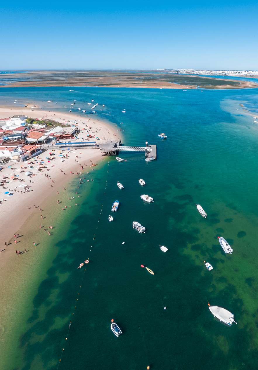 Ria Formosa, no Algarve, com vários barcos a navegar pela água azul da ria, e um aldeamento ao fundo