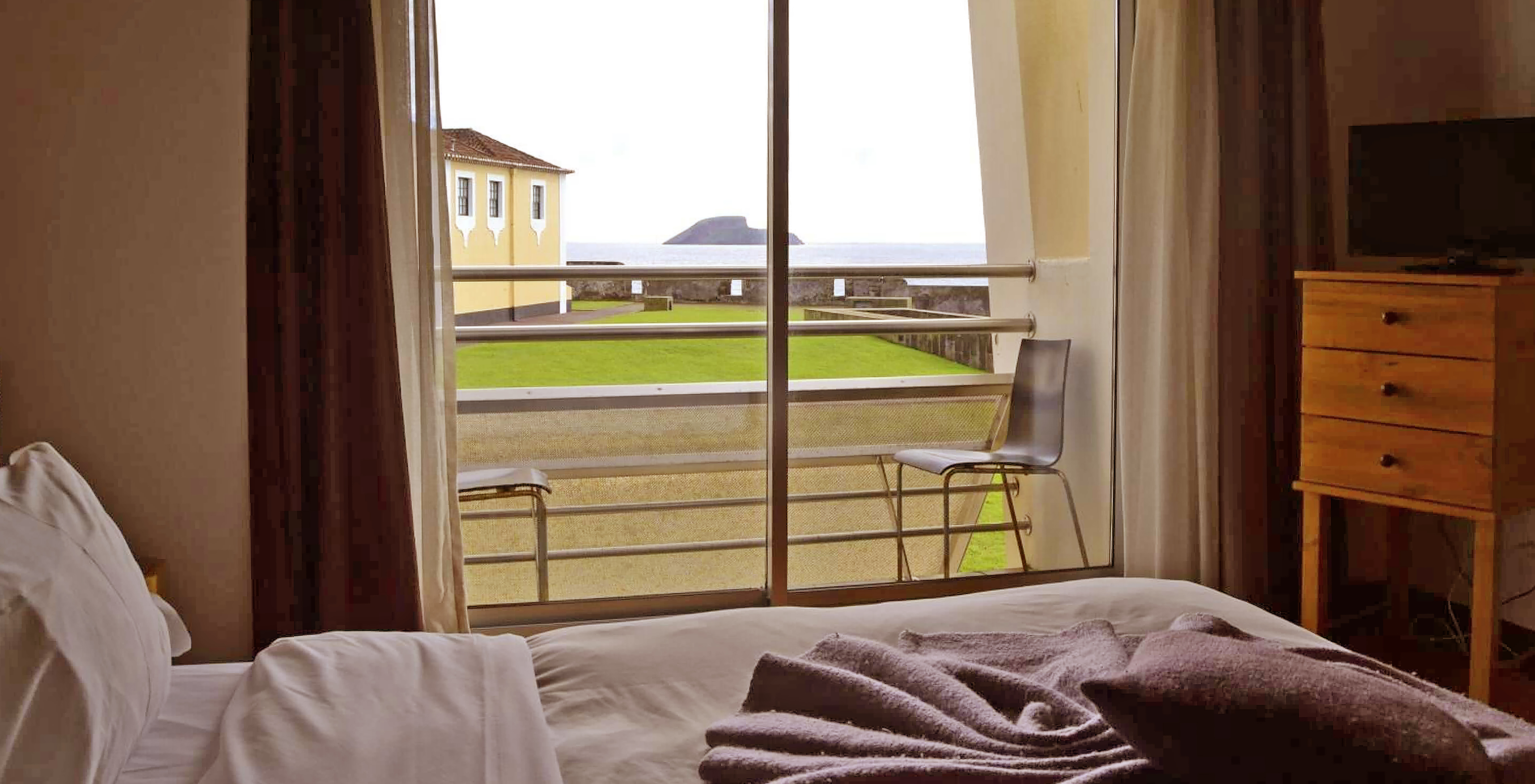 O Quarto Superior da Pousada Forte Angra do Heroísmo tem uma cama, televisão e varanda com duas cadeiras e vista para o mar
