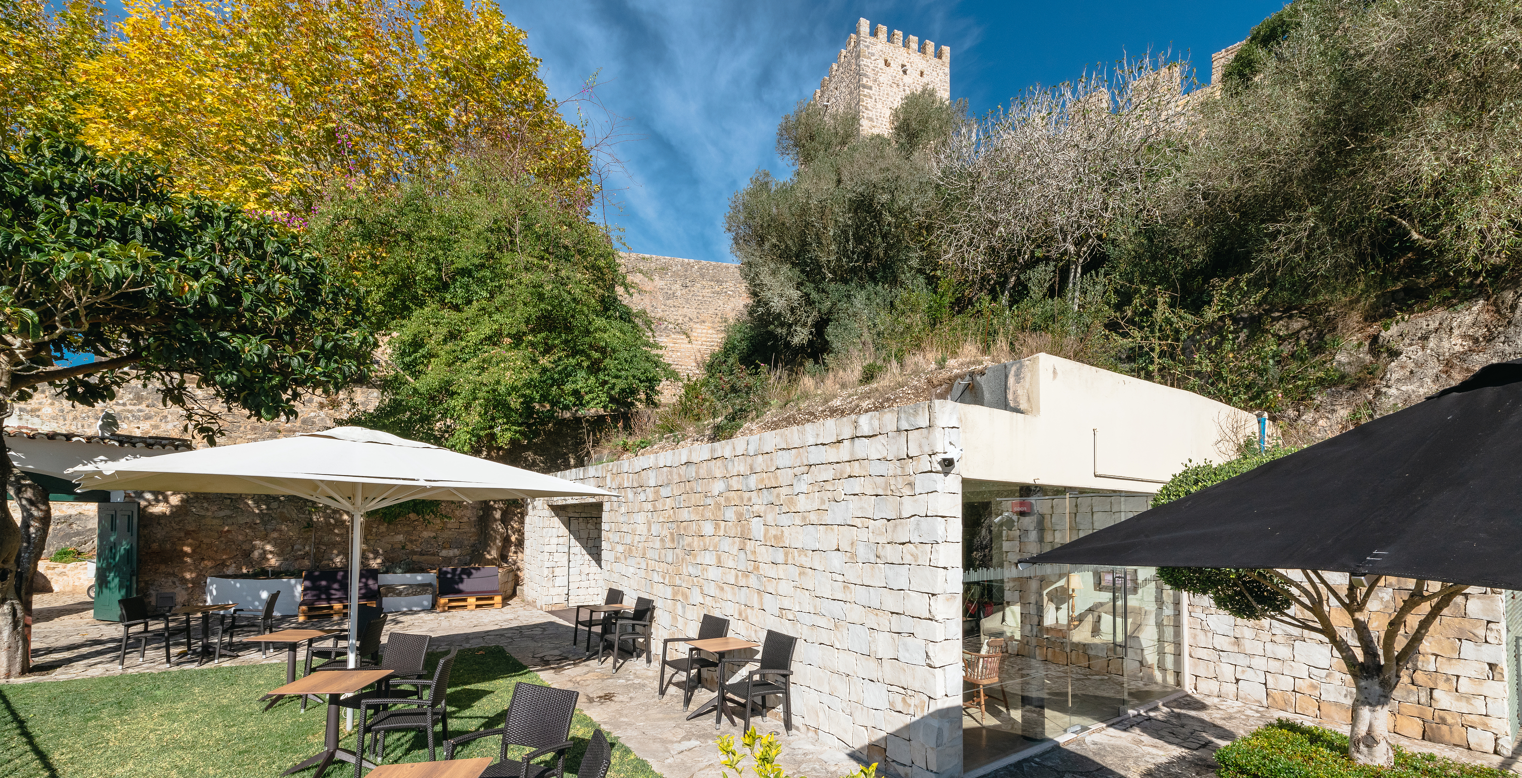 Vista exterior dos jardins da Pousada Castelo Óbidos, com cadeiras e mesas com chapéus para as pessoas usufruírem
