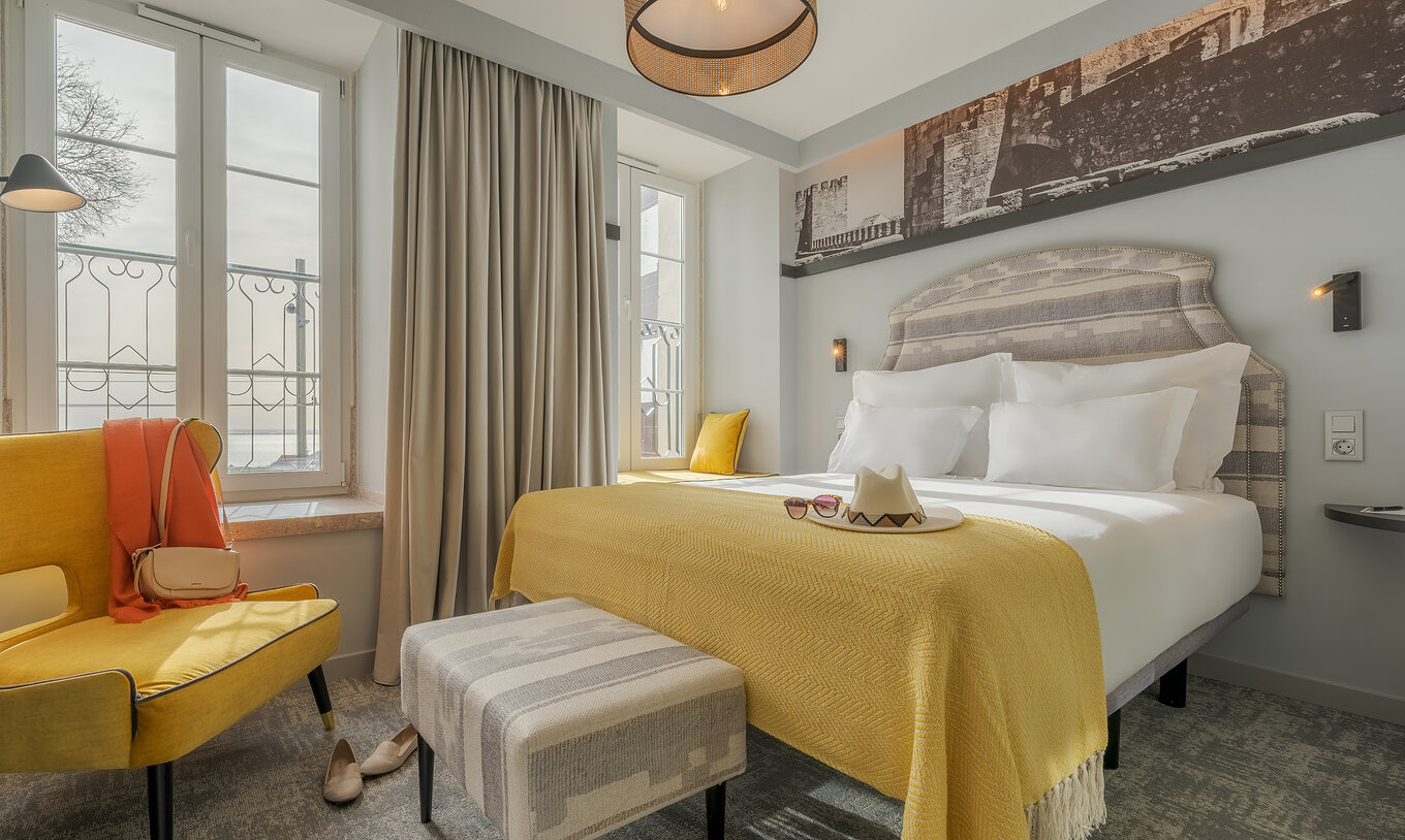 O Superior River View da Pousada Alfama tem uma cama de casal com uma pequena banqueta em frente e um cadeirão amarelo 