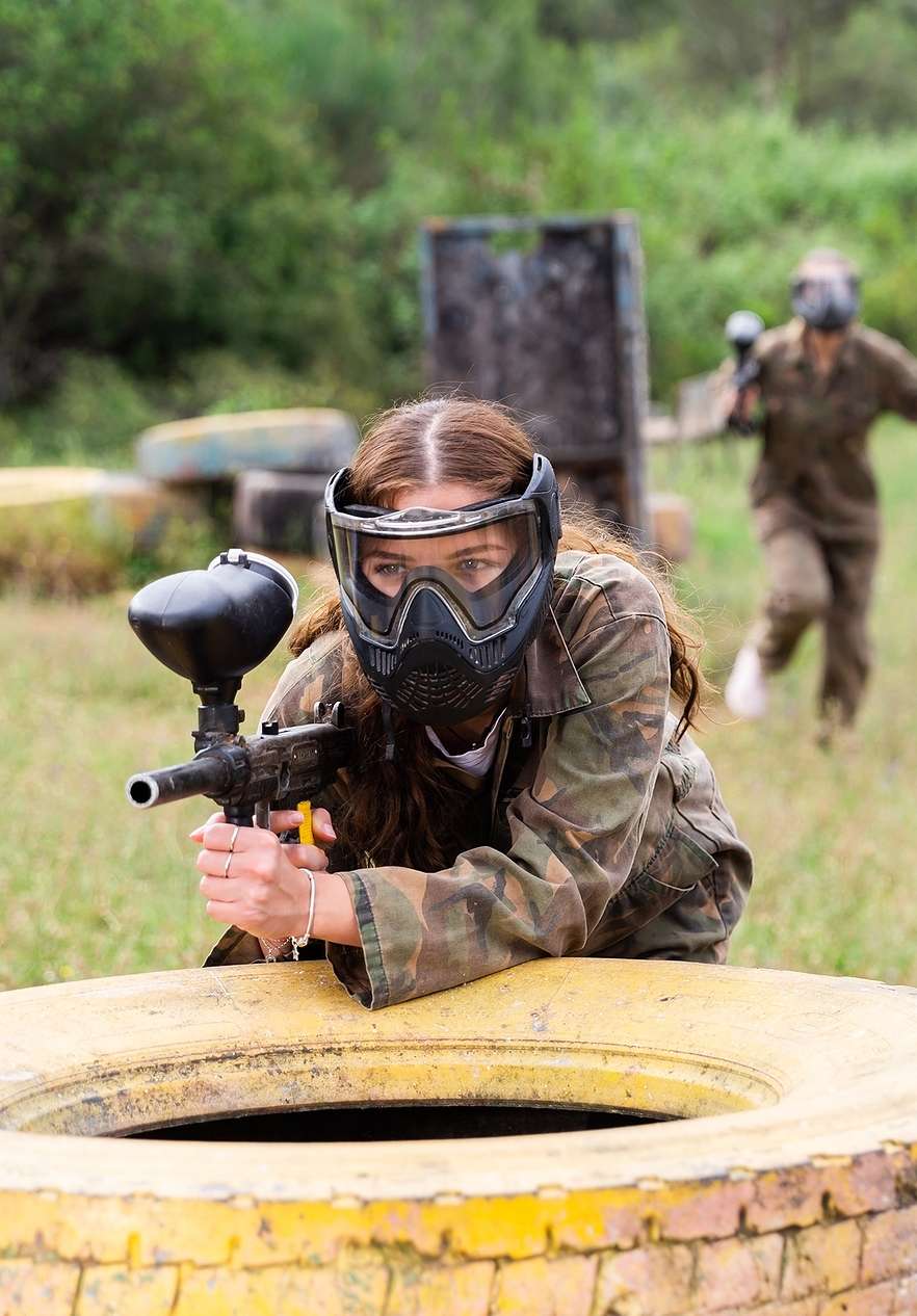 Rapariga com capacete e farda de tropa, com uma arma de paintball, escondida atrás de um pneu amarelo