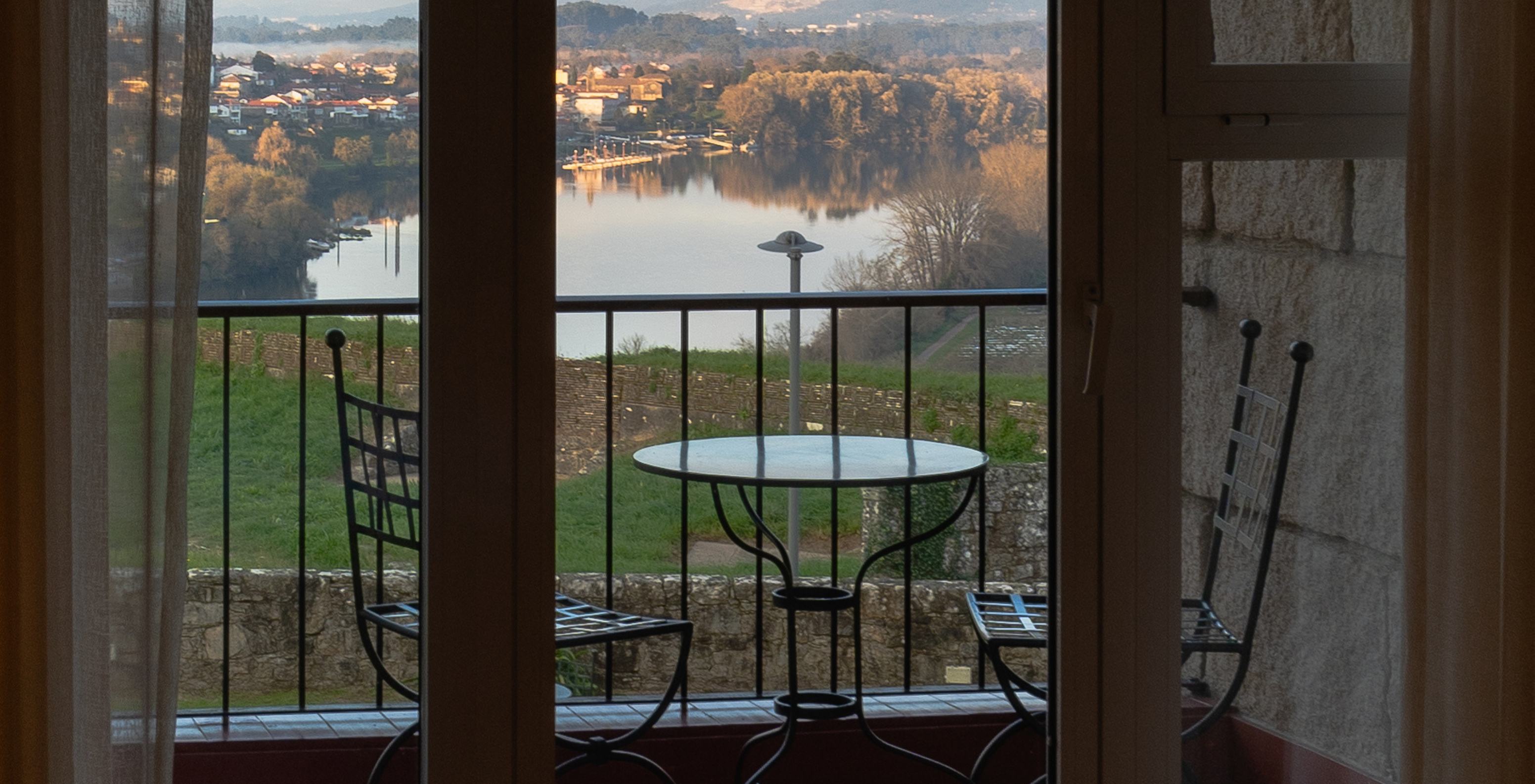 Vista do Pousada Valença, com mesa e cadeiras na varanda e paisagem de rio e colinas ao fundo