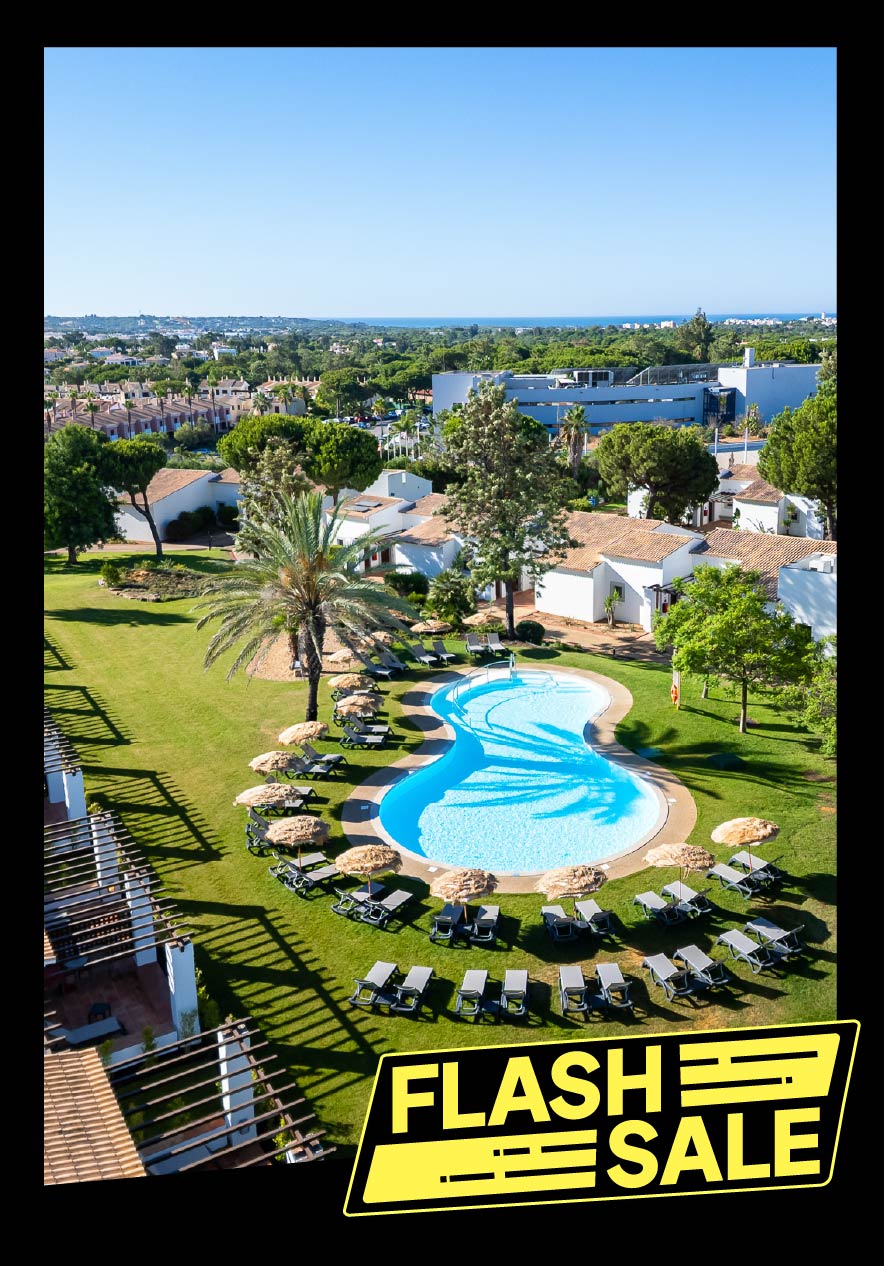 Vista aérea de villas brancas com piscina curva rodeada por relvados, árvores e espreguiçadeiras ao sol