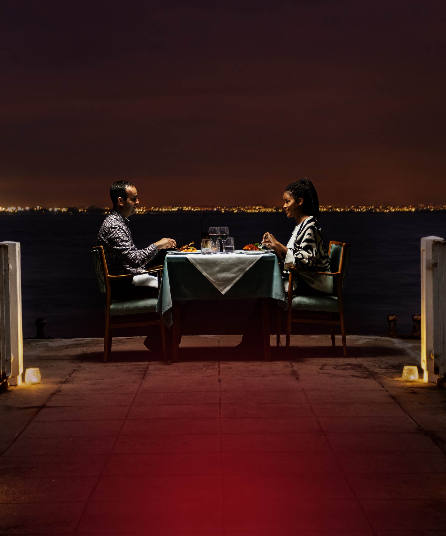 Casal janta numa esplanada junto ao mar à noite, com mesa iluminada e horizonte urbano ao fundo distante
