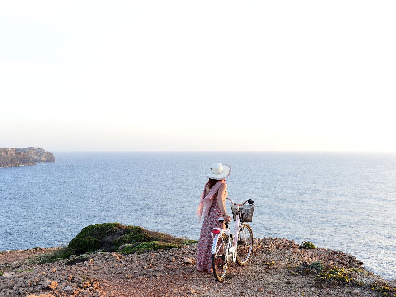 Junge Frau mit Hut und Fahrrad in der Hand, die den Meerblick von einer Klippe in der Algarve genießt