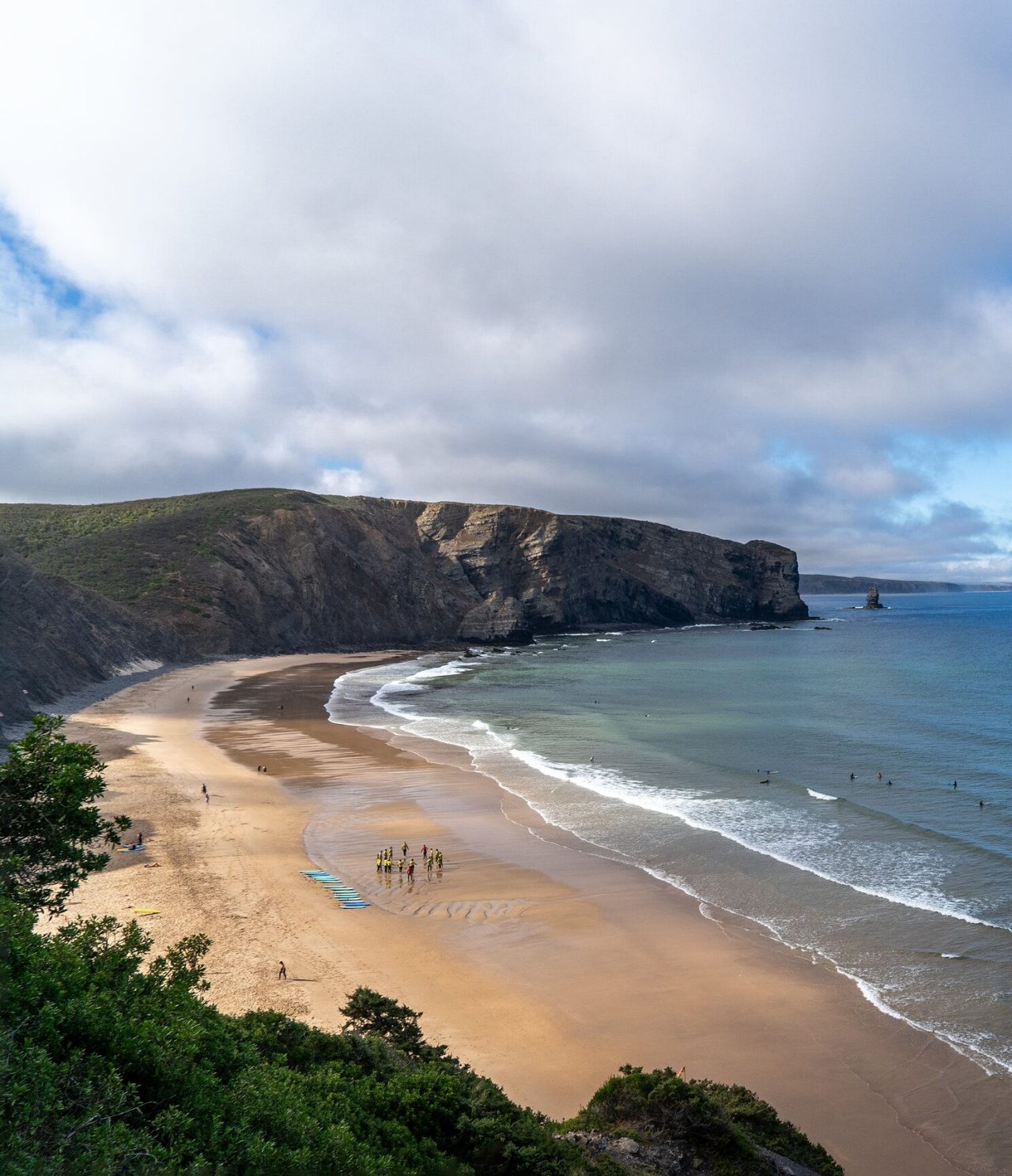 Panoramic view of an extensive beach with rocky cliffs and surfers riding the waves, in the Algarve region