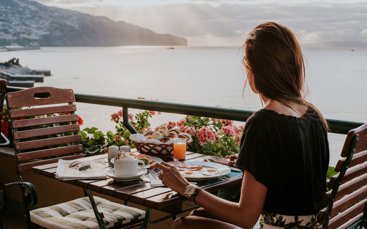 Woman having breakfast on a flower-lined terrace overlooking calm coastal water and distant mountains at sunrise