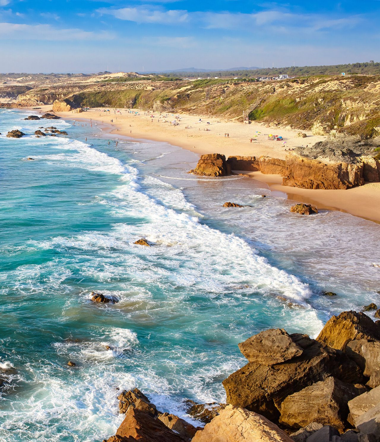 Vista panorámica de la costa alentejana, con una extensa playa de arena dorada, rocas y mar agitado, bajo la luz del sol