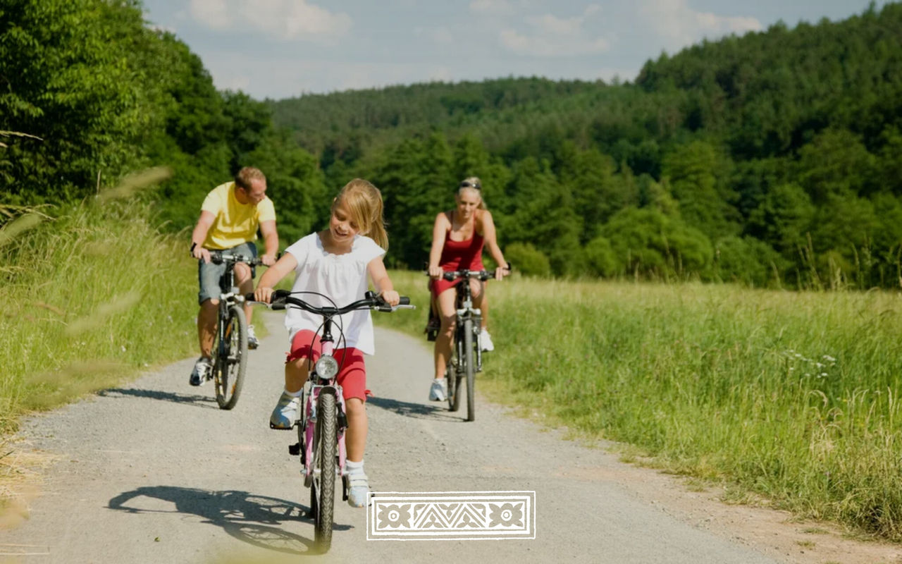 Familia recorriendo en bicicleta un camino rural entre campos verdes y bosque bajo un cielo despejado