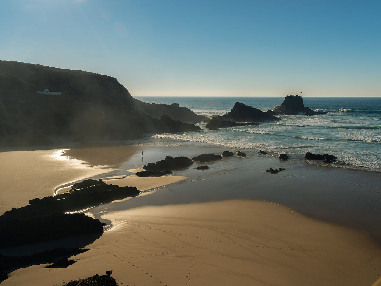 Vue aérienne d'une plage déserte avec des falaises rocheuses, une mer agitée et une personne marchant sous un ciel bleu