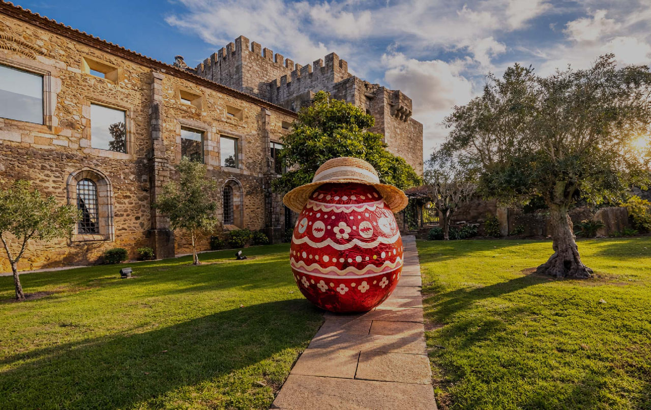 Grande ovo da Páscoa decorado com chapéu de palha em frente a edifício histórico de pedra e jardim verde
