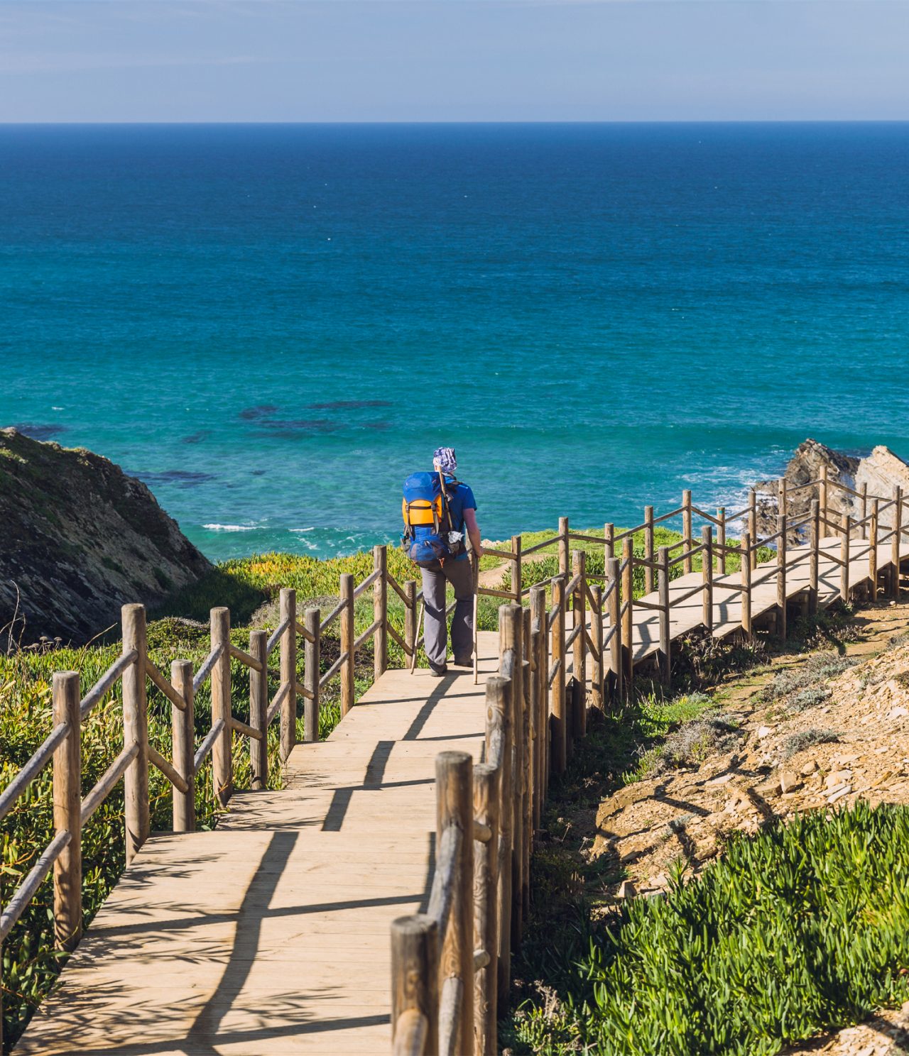 Explore Hotéis no Alentejo com vista para trilho costeiro junto ao mar azul e visitante a caminhar na falésia