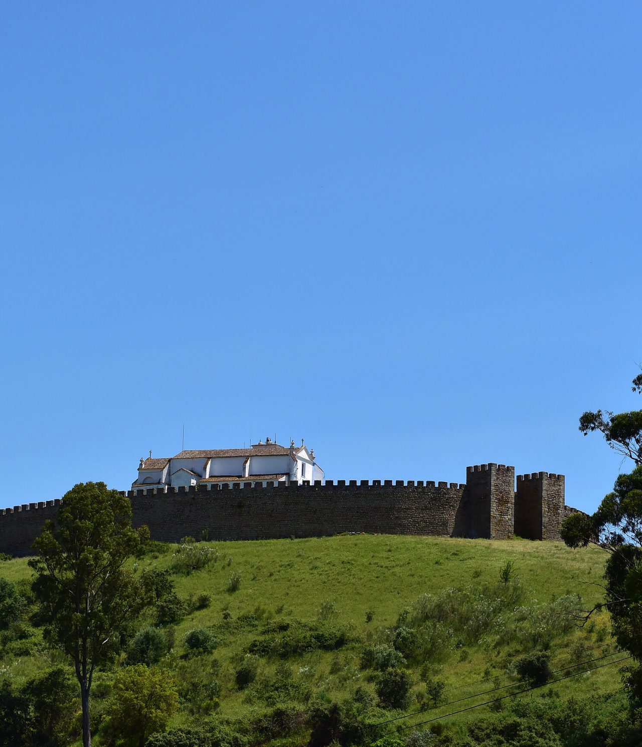 Explore Hotéis no Alentejo com vista para muralhas históricas sobre uma colina verde sob céu azul intenso